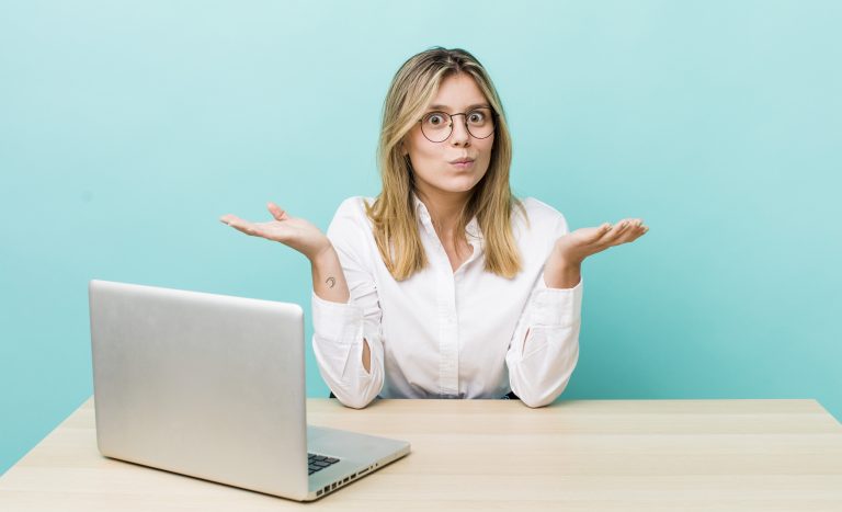 A woman with long blonde hair and glasses sits at a desk with a laptop in front of her. She is wearing a white button-up shirt and has a puzzled expression, with both hands raised and palms up in a questioning gesture.