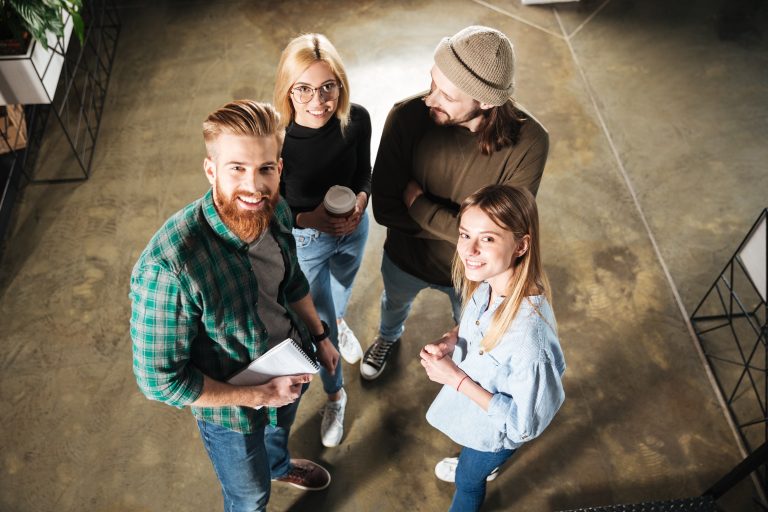 Overhead view of four young adults standing indoors, smiling and talking. One holds a notebook, another holds a coffee cup