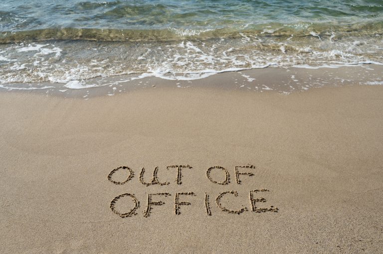 Words 'OUT OF OFFICE' written in the wet sand near the shoreline of a beach, with waves approaching from the sea.