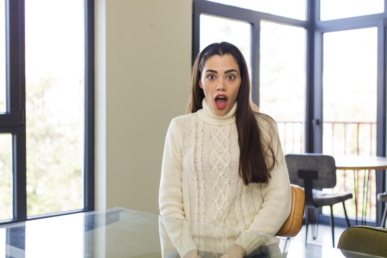 A young woman wearing a white cable-knit sweater sits indoors at a glass table, looking shocked with her mouth open and eyes wide