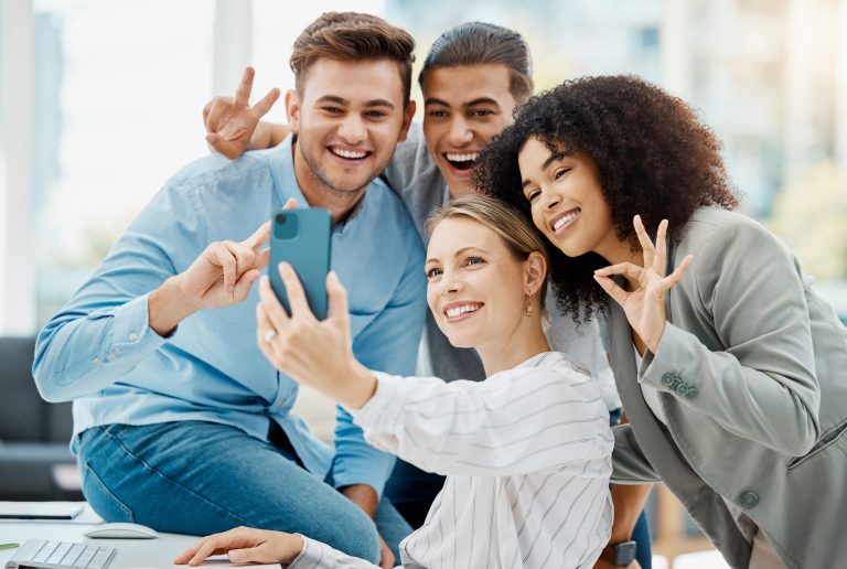 Four cheerful colleagues taking a selfie in an office setting. They are smiling and making playful hand gestures, including peace and OK signs, while sitting together around a desk.