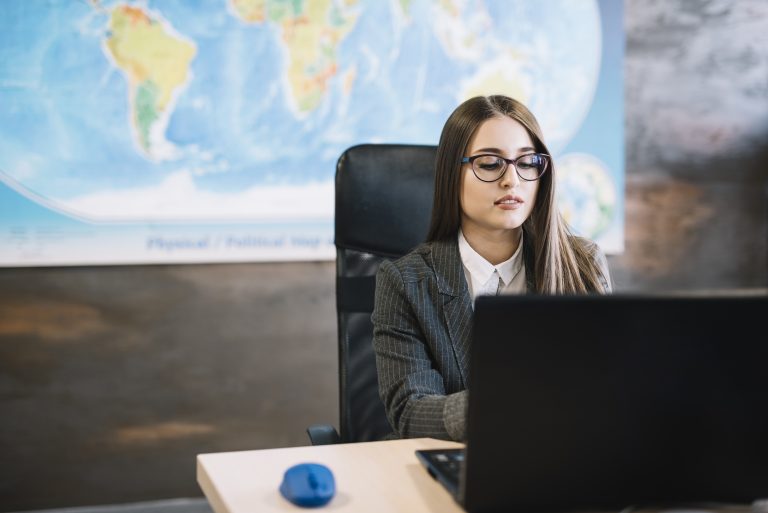 Employers in Europe turn to cross-border hiring to expand their talent pool, survey finds  A young woman wearing glasses and a gray pinstripe suit is sitting at a desk, working on a laptop. There is a world map on the wall behind her and a blue computer mouse on the desk.
