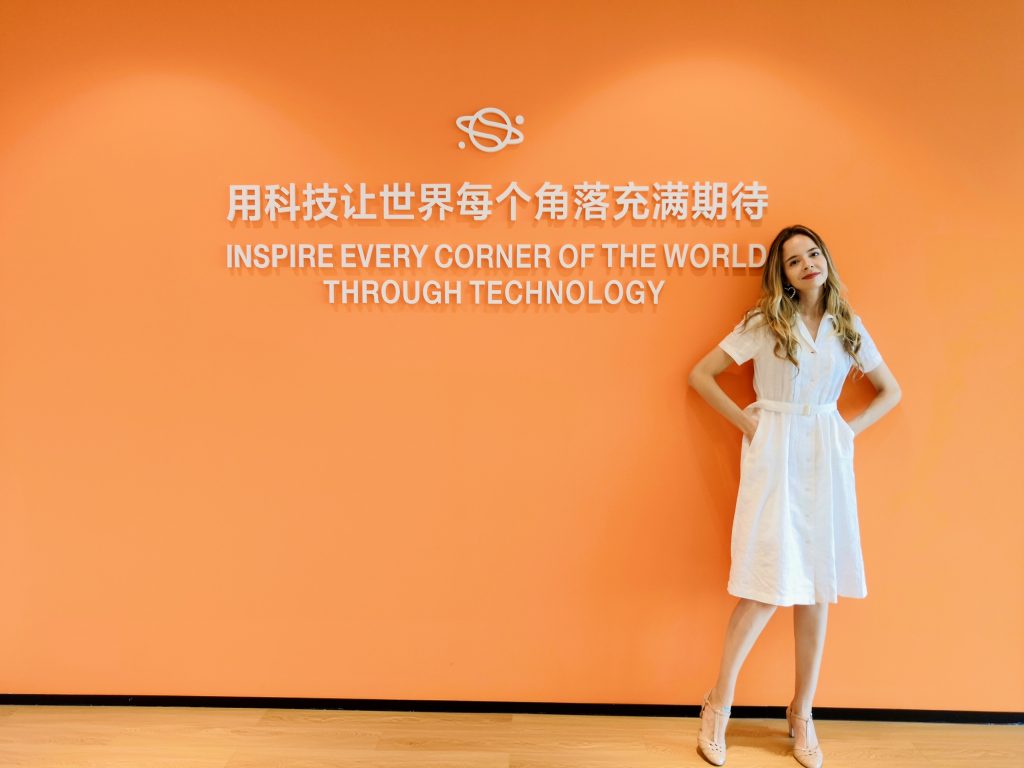 A woman stands against a bright orange wall with white text that reads, 'INSPIRE EVERY CORNER OF THE WORLD THROUGH TECHNOLOGY,' along with Chinese characters. The setting has a modern, professional atmosphere with a wooden floor.