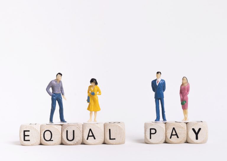 Miniature figurines of two men and two women standing on cubes that spell out the phrase “EQUAL PAY.” The men and women are placed in pairs on either side of the words, symbolizing the concept of gender pay equality.