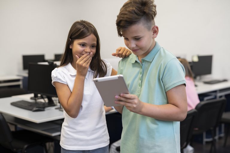 ChatGPT Explains Tech Jobs to a 5-Year-Old (And Accidentally to Ourselves) Two children looking at a tablet together. The boy is holding the tablet while the girl stands beside him, smiling and covering her mouth in amusement.