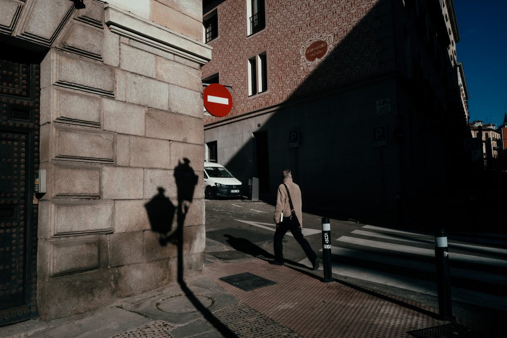A person wearing a beige coat walks across a sunlit street corner in an urban area. Strong shadows from a streetlamp stretch across the stone wall of a historic building. A red “no entry” sign stands above the person, and a white van is parked in the background. Much of the scene is in deep shadow, contrasting with bright patches of sunlight.