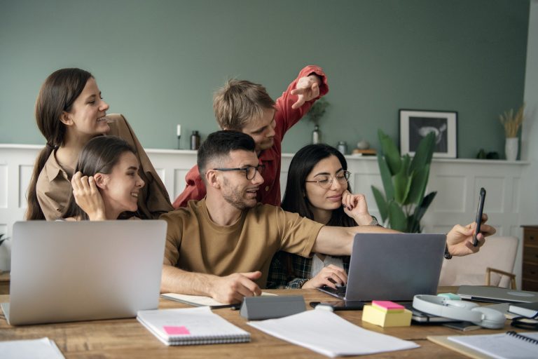 A group of young software developers smiles and takes a selfie while sitting at a table with laptops.
