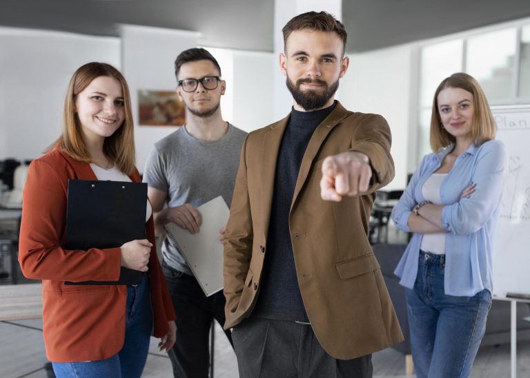 Employer Branding in Tech Recruitment: How To Attract and Win Over Top Talent A tech worker is pointing at the camera while surrounded by three software developers in an office setting. The image represents a technical team in the IT industry