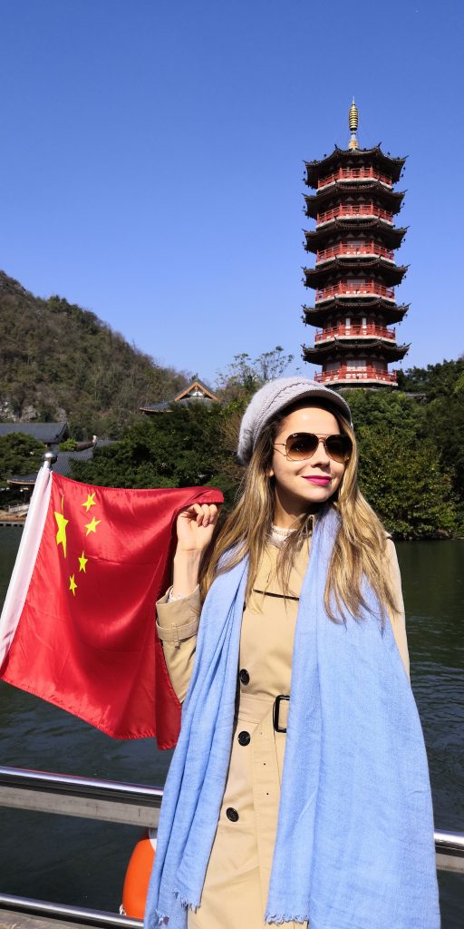 TechTalents Insights' Editor-in-Chief, Gabriela Quintela, during a trip to Guilin in January 2020. She stands on a boat holding a Chinese flag. Behind her, there is a traditional pagoda, surrounded by green trees and mountains. The sky is clear and bright blue.