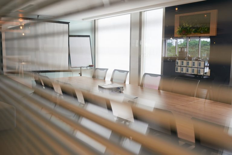 A modern conference room in a tech company seen through partially closed blinds, showing a long meeting table surrounded by mesh office chairs.
