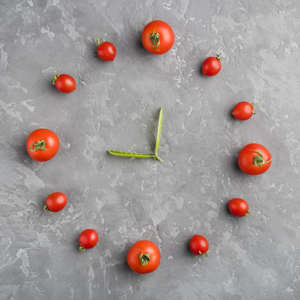 A flat-lay image of various tomatoes arranged in a circle to resemble a clock face. Larger tomatoes mark the 12, 3, 6, and 9 positions, and smaller tomatoes fill in the rest of the hours. Two green leaves in the center act as the clock’s hands, all set on a textured gray surface.