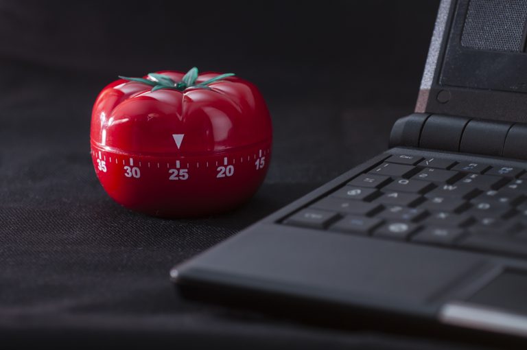 A red tomato-shaped kitchen timer set to 25 minutes sits on a black surface next to a partially visible black laptop keyboard, suggesting use of the Pomodoro Technique for time management. The image represents the Pomodoro Technique for software developers