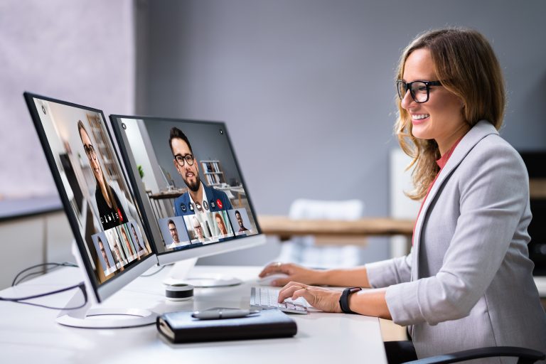 Remote Employee Management: Best Practices From Tech CEOs A female tech worker is attending a videoconference while sitting at a desk with two monitors displaying her remote software developer colleagues