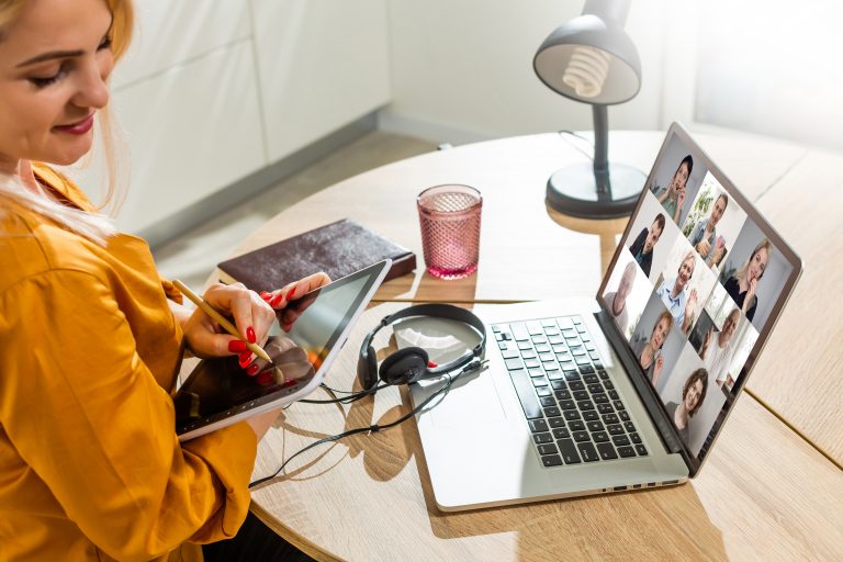 A female tech worker participates in a virtual meeting from home. She is smiling and using a stylus on a tablet while a laptop in front of her displays a video call with nine software developers/tech workers. The setting is bright and modern, suggesting a remote work environment.