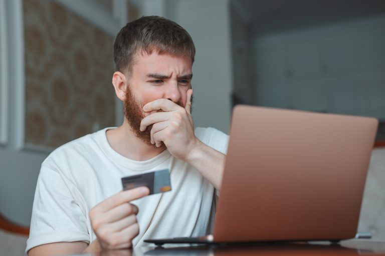 A concerned tech worker sitting at a desk, holding a credit card while looking at a laptop screen, with his hand covering his mouth, suggesting worry about an online payment required by a freelancing platform.