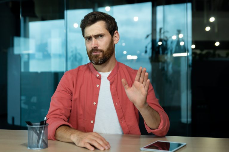 A developer sitting at a desk raises his hand in a stopping gesture, appearing to decline a job offer during an interview.