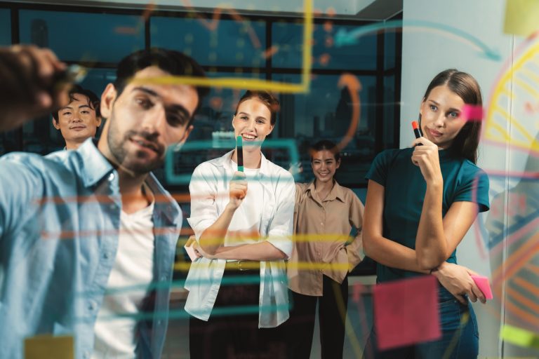 Gen Z More Willing Than Older Workers To Accept Fully In-Office Work, Survey Shows Four young tech professionals collaborating in a modern office, writing colorful notes and diagrams on a glass board with markers while discussing ideas.