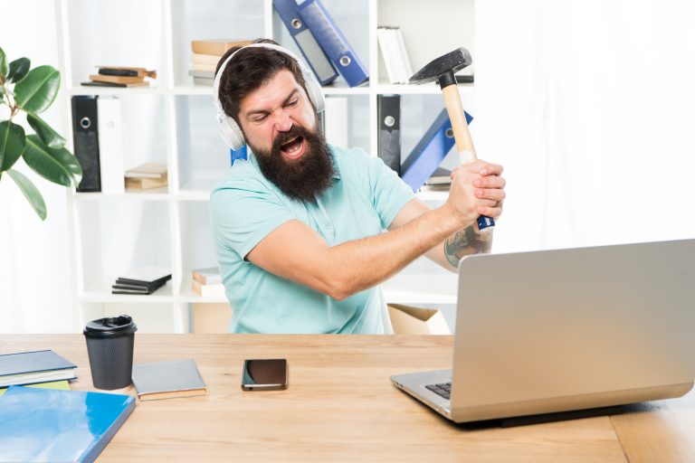 Frustrated tech worker at a desk raises a hammer toward a laptop, reacting angrily as if fed up with misleading or disappointing job perks.