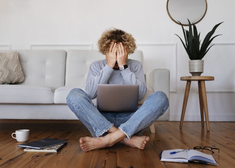 A tech worker sitting on the floor at home with a laptop, covering their face in stress, illustrating burnout and isolation caused by prolonged remote work