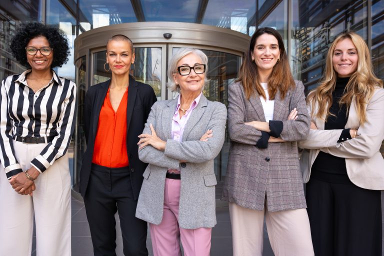 Five professional female tech workers of different ages and ethnic backgrounds stand confidently outside a modern office building. The group represents workplace diversity and the implementation of the EU Pay Transparency Directive