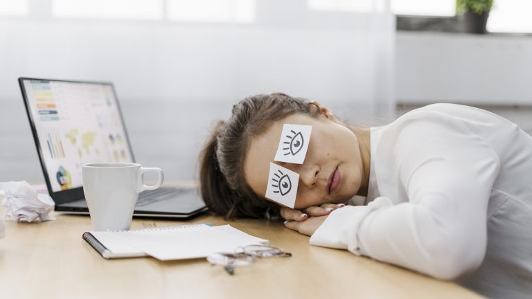 A tired office worker rests her head on a desk beside a laptop, coffee mug, notebook, and glasses, with sticky notes showing drawn eyes covering her closed eyes to symbolize exhaustion.