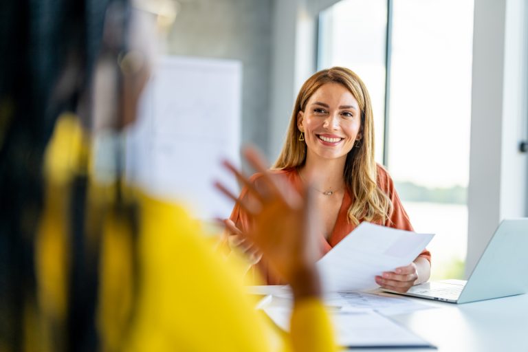 Smiling woman during a job interview holding documents, gesturing while talking with the interviewee — representing instinct during tech recruitment
