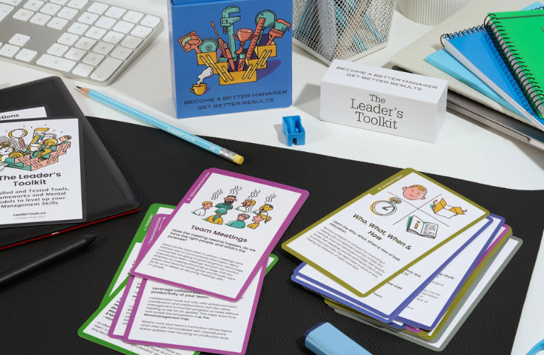 A desktop workspace featuring The Leader’s Toolkit management card game. Color-coded decks of illustrated leadership prompt cards are spread across a black desk mat, alongside a tablet displaying the toolkit cover, a keyboard, a pencil, notebooks, and office supplies. Branded packaging and a box labeled “The Leader’s Toolkit” sit in the background, emphasizing a professional leadership development resource.