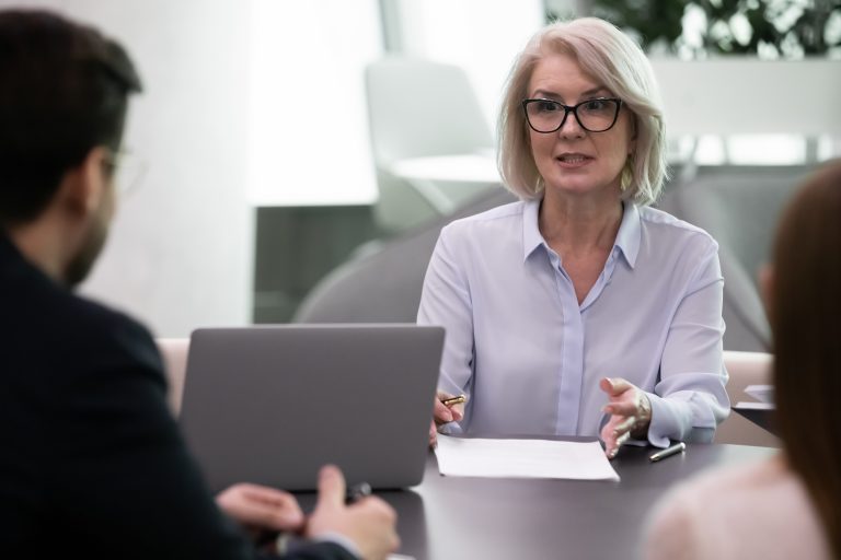 Am I Allowed To Ask a Candidate’s Age? An older tech worker with glasses speaks with two people across an office table, with an open laptop and paperwork between them.