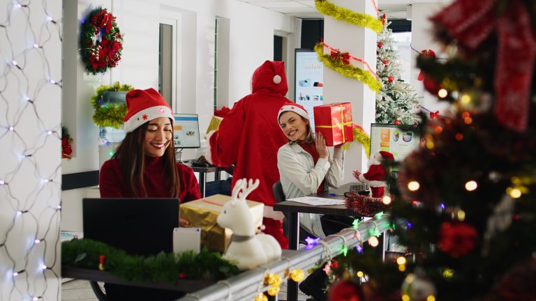 Tech workers in Santa hats exchange gifts with a colleague dressed as Santa in an office decorated with Christmas trees, garlands, and string lights.
