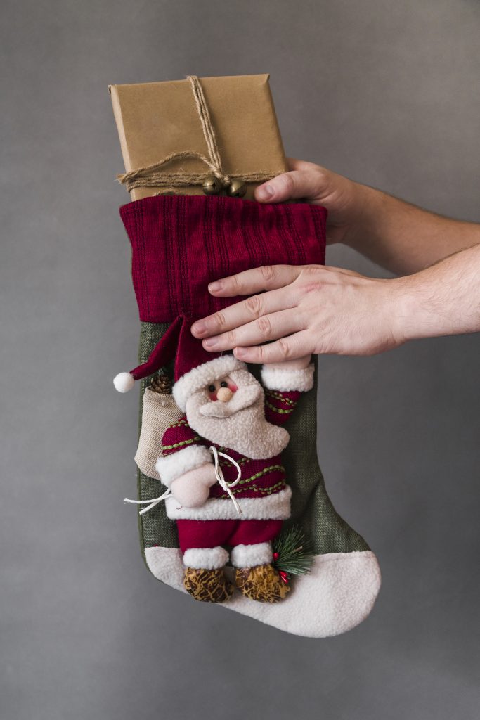 Hands pulling a gift wrapped in brown paper and tied with twine and bells from a Christmas stocking decorated with Santa Claus